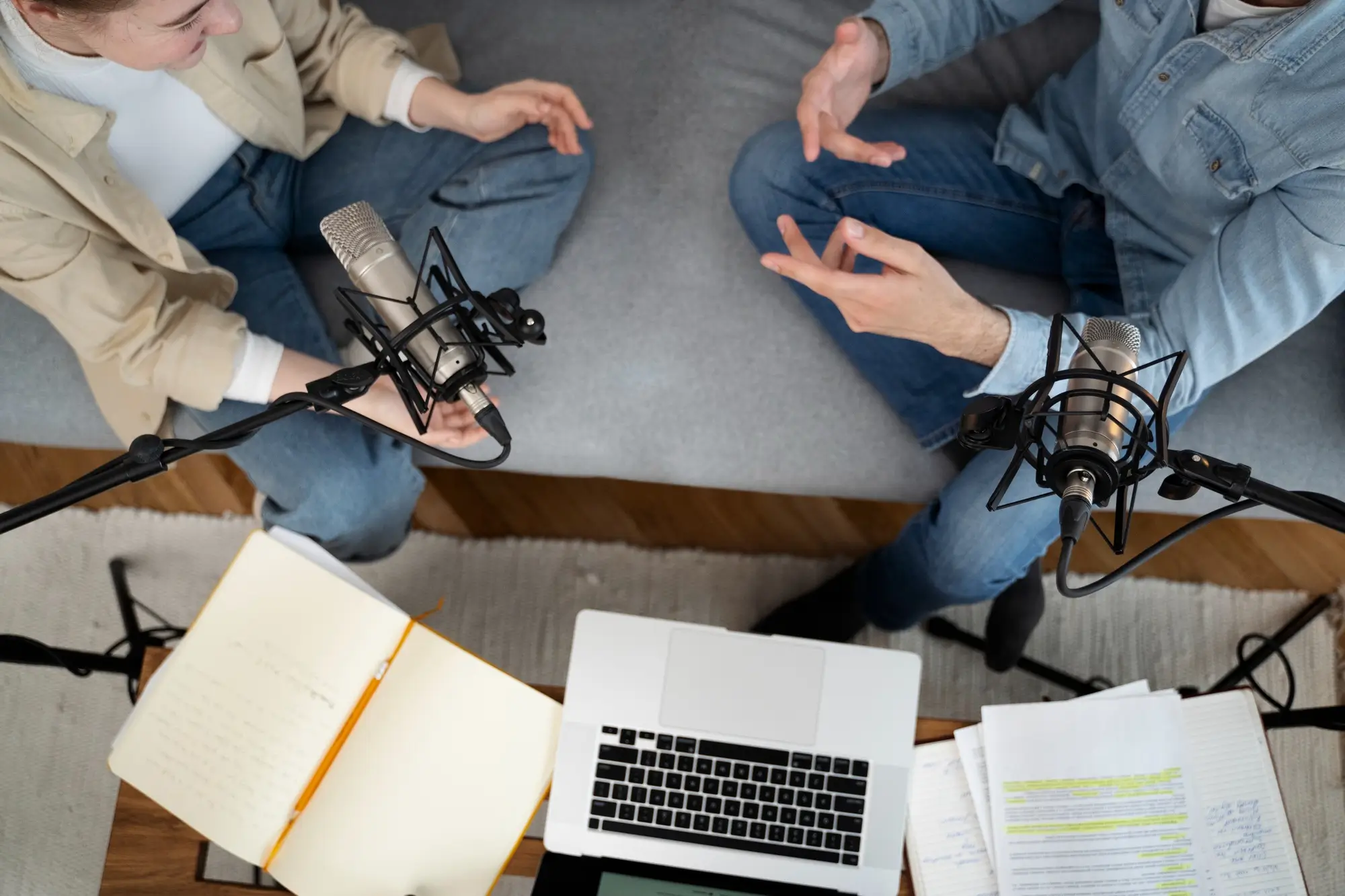 Two people sat on a sofa with microphones, a laptop and paper - making a podcast