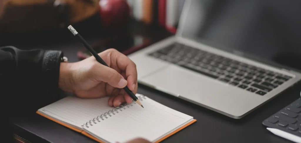 Close up of a man's hand writing in a notebook with a laptop in the background