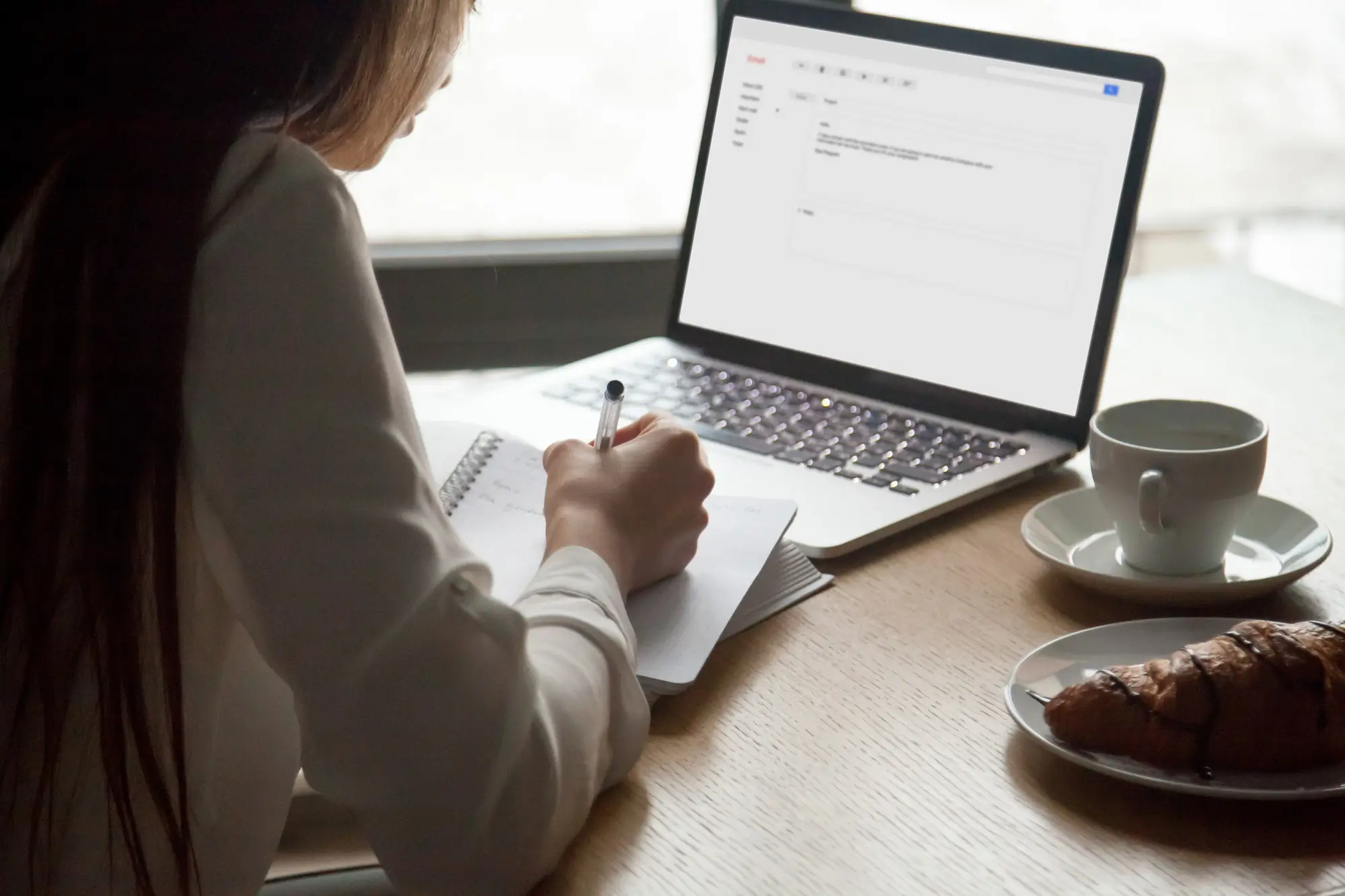A woman writing in a notebook with a laptop in front of her